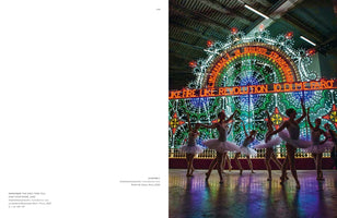 An art installation view showing several ballerinas in white tutus performing in front of a massive, ornate light structure. The backdrop glows with intricate patterns of green, blue, and white lights and features red neon text, including the phrase LIKE FIRE LIKE REVOLUTION. The colorful lights create vibrant reflections on the floor around the dancers.