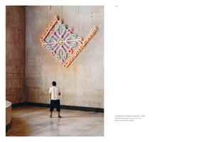 A page from a book featuring a photograph on the left and text on the right. The photo shows a person from behind, standing on a polished stone floor and looking up at a large, diamond-shaped neon art installation on a stone wall. The colorful artwork has the words A BLEND OF COMMON HUMANITY in orange neon along its edges.