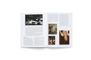 An overhead shot of an open book on a white surface. The two-page spread discusses the art market, with the left page showing a photo of a modern auction room. The right page displays text and three images of paintings: one abstract floral, one of a figure in masks, and one of an upside-down woman.