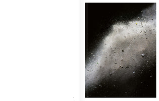 A two-page book spread with a blank white page on the left. The right page features a dramatic, full-bleed photograph of a cloud of white and grey dust, powder, and small rocks exploding diagonally against a stark black background.