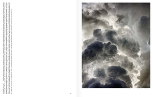 An open art book spread. The right page features a full-bleed, vertical photograph of dense, turbulent clouds in dramatic shades of dark grey and bright white, illuminated from behind. The left page contains a column of text that identifies the image as a work by Andreas Gefeller, explaining that the clouds are industrial water vapor emissions from a power plant.