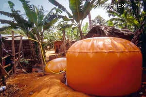 An outdoor photo of a rural village in a tropical climate, with two large, orange, flexible biogas digesters sitting on red dirt ground amidst banana trees and simple huts. A yellow hose connects to one of the digesters, and a young child stands to the left.