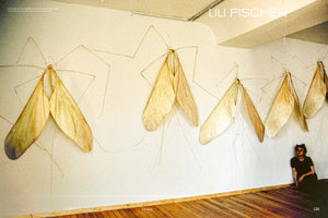 An indoor photograph shows artist Lili Fischer sitting on the wooden floor of a gallery, next to her installation. On the white wall, a row of large sculptures resembling giant moths or mayflies is displayed. The artworks have delicate, translucent wings in shades of cream and brown, with thin wire bodies and antennae.