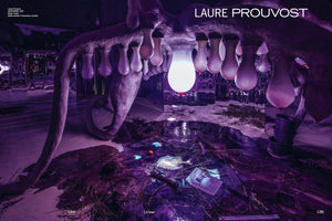 A surreal art installation by Laure Prouvost, awash in purple light. A large, fleshy, udder-like sculpture hangs overhead, with its teats made of glowing glass vessels of various sizes. The floor beneath is a messy pool of dark liquid littered with branches, debris, an orange, and an open book.