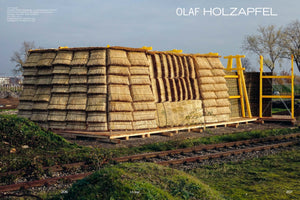 An outdoor art installation by Olaf Holzapfel featuring a long, barn-like structure made of tiered layers of stacked rye straw. The golden-brown thatched building sits on a wooden platform next to rusty train tracks in a grassy field. A bright yellow timber framework is visible at one end under an overcast sky.