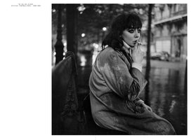 A black and white photograph by Ed van der Elsken showing a young woman, Vali Myers, sitting on a bench on a wet Paris street at night, circa 1950-1954. She has dark, messy hair, heavy eyeliner, and a pensive expression as she brings a cigarette to her lips. She wears a large coat over a turtleneck, and the city lights are blurred in the background.