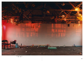A wide, atmospheric shot of the aftermath of a party in a large warehouse. The concrete floor is strewn with litter, empty bottles, and crumpled debris. On the left, a few chairs are clustered together. Above, strings of fairy lights and bunches of flowers hang from the high, exposed ceiling trusses. The back wall is illuminated by a reddish-orange glow, which casts large shadows of windows across its surface.