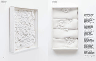 A page from an art book displaying two all-white, three-dimensional wall sculptures by Ferdinand Spindel. The piece on the left, from 1974, is a single, turbulent mass of foam-like, bubbly textures inside a white frame. The piece on the right, from 1977, features four stacked, soft, horizontal forms with smaller lumpy clusters on their surfaces, also in a white frame. A block of German text is next to the artwork on the right.