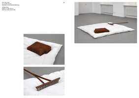 A page from a catalog showing an art installation. The main photo shows a white duvet spread on the grey floor of a white room. A rusty, rectangular cast-iron object shaped like a pillow and a rusty metal rake lie on top of the duvet. Two smaller photos below show close-ups of the cast-iron pillow and the rake.