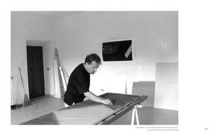 A black-and-white photograph from 1990 shows artist Jan Jedlička working in his studio. He wears a dark shirt and leans over a long workbench, focused on rubbing the surface of a large mezzotint plate. The studio background includes a dark door, a broom, rolled canvases, and an abstract print on the wall.