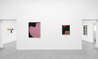 A wide shot of a minimalist art gallery with white walls and a concrete floor. Four abstract paintings are displayed; two on the central wall feature bold shapes in pink, red, and brown on black backgrounds, while two others, one geometric and colorful and one with a black and white pattern, are visible through doorways on either side.