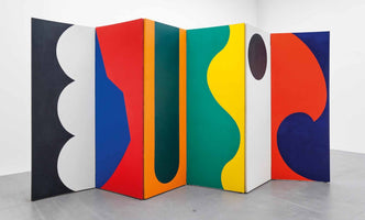 A freestanding, multi-paneled sculpture resembling a folding screen, set in a white gallery. Each panel is painted with large, abstract shapes in bold, solid colors, including red, blue, yellow, green, black, and white. The designs feature a mix of organic curves, scalloped edges, and hard-edged geometric forms.