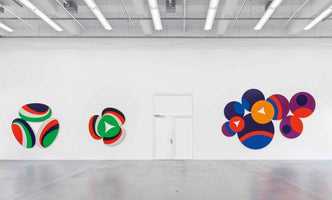 An interior shot of a modern art gallery with a high industrial ceiling, white walls, and a concrete floor. Three colorful, abstract geometric artworks hang on the main wall. On the left are two smaller pieces made of layered ovals with red, green, blue, and white stripes. On the right is a larger composition of overlapping circles in purple, blue, orange, and red. A white door is positioned in the center of the wall between the artworks.