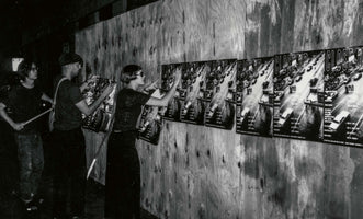 A grainy, black and white photograph of three young people putting up posters on a plywood wall at night. A woman with a bob haircut and sunglasses presses a poster onto the wall, joining a row of identical posters that feature an image of a highway and the words CLOSE-UP. Two men stand beside her, one holding more posters.