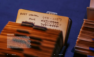 A close-up, angled shot of a vintage Rolodex card. The yellowed card has handwritten entries in black ink for Andy Warhol, with phone numbers for his Home and Factory. Some of the numbers are crossed out. The card sits in an index against a dark blue background.