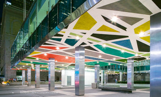 A wide, low-angle shot of the ground floor exterior of the Lever House building at night. The ceiling of the open-air plaza is covered in a vibrant, colorful mural of abstract geometric shapes in yellow, red, green, blue, and black, separated by thick white lines and lit by spotlights. The ceiling is supported by numerous large, square, reflective stainless steel columns.