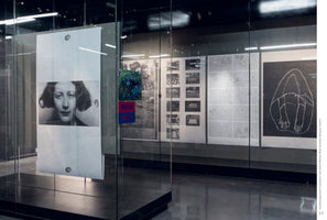 An interior shot of a modern art gallery showing several pieces of art behind glass. In the foreground, a large white sheet hangs in a glass case, featuring a black-and-white photographic portrait of a woman. In the background, a wall displays a series of other black-and-white artworks, including photographs and line drawings.