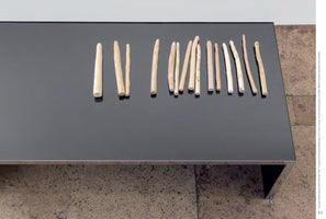 A high-angle photograph of a minimalist art installation. Fifteen carved wooden asparagus spears of varying sizes are arranged in a row on a sleek, modern table with a black, reflective top. The table is placed on a rough, textured concrete floor.