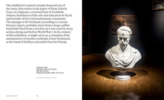 A two-page spread from an exhibition catalog. The right page shows a photograph of a large, weathered plaster bust of Archduke Johann of Austria, displayed in a dark gallery. The bust is spotlit from above, highlighting a distinct bullet hole in its forehead, and rests on a polished wooden floor. The left page contains text describing the bust and the historical context of its damage.
