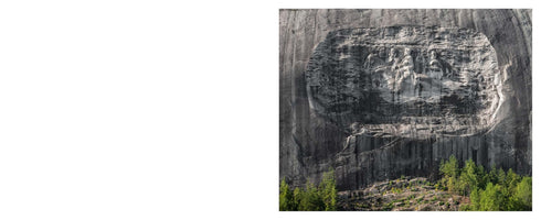 A wide shot of the Confederate Memorial Carving on the granite face of Stone Mountain, Georgia. The massive bas-relief sculpture depicts three Confederate leaders on horseback. A line of green trees is visible at the base of the mountain.
