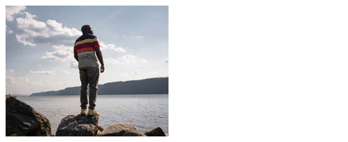 A Black man seen from behind, wearing a colorful striped t-shirt and jeans, stands on a large rock at the edge of a wide body of water. He looks out across the water towards a tree-covered shoreline in the distance under a blue sky with scattered white clouds.