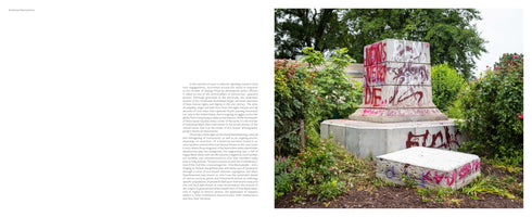 A two-page spread from a book titled American Monuments. The left page contains text, and the right page shows a photograph of a stone monument pedestal in a grassy area with trees. The statue is gone, and the pedestal is covered in red and black spray-painted graffiti. A separate, graffiti-covered block of stone rests on the ground in front of it.