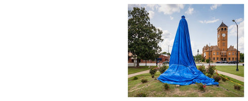 A tall monument on a grassy mound is completely covered by a large, bright blue tarp. In the background, a historic brick courthouse with a clock tower stands across the street under a partly cloudy sky.