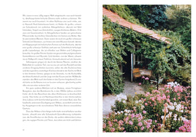 An open book with German text on the left page and a full-page color photograph on the right. The photo shows a rustic, single-story house with a red-tiled roof, almost completely covered in lush green vines. In the foreground, a vibrant garden bed is filled with pink and white flowers, and a small table with chairs sits on a patio near the open doorway.