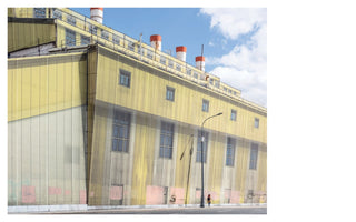 A low-angle photograph of a massive, yellow industrial building, its facade covered by a semi-transparent scrim that partially obscures the windows and walls beneath. Four red-and-white-topped smokestacks rise from the roof against a blue sky with white clouds. On the sidewalk in the foreground, a single person in a red hat walks past a streetlight, dwarfed by the building's immense scale.