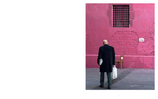A street photograph, presented on the right side of a white page, shows a bald man in a long black coat from behind. He stands on a sidewalk, holding a newspaper and a white plastic bag, facing a vibrant, weathered pink wall with a barred window and a lone wooden chair resting against it.
