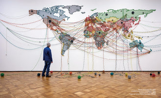 An installation view of a large world map made from colorful woven material on a white gallery wall. A person in a blue jacket stands looking at the map. Multicolored threads crisscross the globe, connecting various points, and then drop down to the parquet floor, ending in scattered balls of yarn.