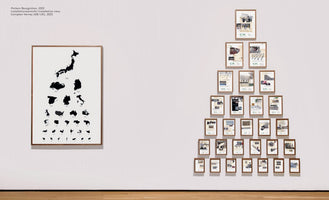 An installation view of two artworks on a white gallery wall. On the left, a large vertical frame contains an artwork resembling an eye chart, but with black silhouettes of different countries that decrease in size. On the right, many smaller framed artworks are arranged in a large triangular formation.