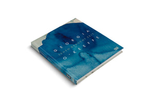 An angled, top-down shot of a square hardcover book on a white background. The cover features a deep blue watercolor design with white, spaced-out text reading GEORGIA O'KEEFFE, SEHEN BRAUCHT ZEIT. The spine is visible on the left.