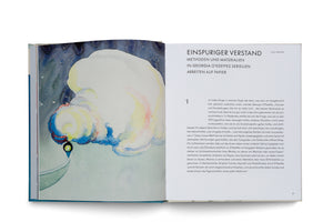 An overhead shot of an open book on a white surface. The left page is a full-color abstract watercolor painting of a puffy, white and yellow cloud-like form against a dark blue sky. The right page contains text in German about the artist Georgia O'Keeffe's work on paper.