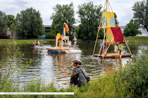 A lively outdoor scene at a park lake where people enjoy a sunny day on creative, homemade rafts. In the foreground, a man and a young boy sit on the grassy bank watching the activity. One raft features a large painted face, while another has a tall wooden A-frame with colorful fabric sails.