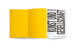 A book with a vibrant yellow cover flap is open on a white surface. The right-hand title page is white and features bold, black, vertical text in German reading KUNST UND GESELLSCHAFT, with the years 1972 2022 2012 printed above. The design is minimalist and graphic.