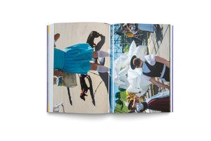 An open photobook on a white surface. The two-page spread features color photographs turned sideways. The left page shows a person in a blue dress and white socks walking two small black dachshunds. The right page shows people in traditional Bavarian attire at an outdoor festival.