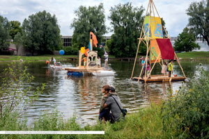An outdoor photo of a man and a boy sitting on a grassy bank, looking out at a lake where several people, including children, are on large, colorful homemade rafts. One raft features a tall wooden A-frame with bright sails, while another is decorated with a large, carved face. The scene is set in a park on a cloudy day.