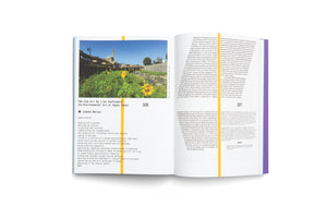 An overhead shot of an open book on a white background. The left page features a large photograph of a city with a river and a mosque, with sunflowers in the foreground, under which is the title We Can All Be Like Sunflowers: An Environmental Art of Agnes Denes. The right page is filled with columns of text.