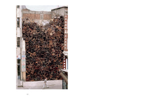 An outdoor art installation featuring a towering, chaotic mass of hundreds of wooden chairs packed tightly into the empty space between two city buildings. A person walks on the sidewalk in the foreground, dwarfed by the scale of the structure.