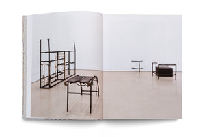 A photograph of an open book displaying a two-page spread of a modern art exhibition in a minimalist gallery with white walls and a light floor, featuring several sparse, industrial-style metal sculptures.