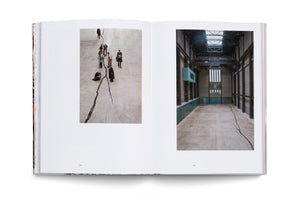 An open art book on a white surface shows a two-page spread. The left page has a high-angle photo of people looking down at a large, jagged crack in a concrete floor. The right page shows a wider photo of the same crack running through the vast, empty, industrial-style gallery hall.