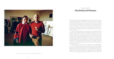 A page from the book My Photos of Picasso by Edward Quinn. The left page displays a color photograph of Pablo Picasso in a red shirt and patterned pants, standing next to his wife Jacqueline Roque, who wears a red poncho. They are in his studio, surrounded by paintings, in Mougins, 1962. The right page contains a block of text.