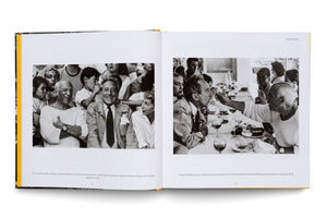 An open book showing a two-page spread with black-and-white photographs from 1955. The left photo shows Pablo Picasso and Jean Cocteau smiling in a crowd. The right photo shows Picasso feeding Cocteau a bite of food at a restaurant table surrounded by friends.