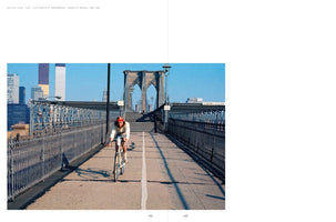 A 1971 color photograph of a man in a red cap riding a bicycle on the pedestrian walkway of the Brooklyn Bridge. In the background, the stone arches of the bridge and the Manhattan skyline, including the original World Trade Center Twin Towers, are visible against a clear blue sky.