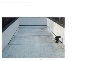 A book spread showing a photograph of a minimalist art installation in an empty, rectangular concrete swimming pool with white walls. A thick, continuous black line runs along the walls and across the floor, forming an inner rectangle. In the far right corner, a person in dark clothing is crouched down, interacting with the line.