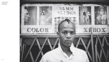 A black and white photograph of a young Jean-Michel Basquiat in New York, 1980. He has a mohawk hairstyle and looks directly at the camera with a serious expression. He stands in front of a storefront with a metal accordion gate and a sign above it with stenciled lettering for COLOR, 35 MM SLIDES, and XEROX.