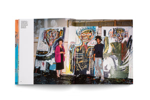 A photograph in an open book shows artist Jean-Michel Basquiat and gallerist Annina Nosei standing in his studio in 1982. Basquiat, on the right, wears paint-splattered clothes, while Nosei wears a black dress and a pink cardigan. They stand on a paint-covered floor in front of three large, colorful neo-expressionist paintings of skeletal figures.