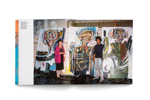A photograph from a book shows gallerist Annina Nosei and artist Jean-Michel Basquiat standing in a messy studio in 1982. Nosei, a white woman in a pink cardigan, stands on the left. Basquiat, a young Black man in paint-splattered clothes, stands on the right. They are in front of three of his large, vibrant, neo-expressionist paintings of skeletal figures. The floor is covered in dark, wet-looking paint.