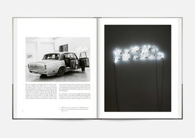 An open art book displaying a two-page spread. The left page has a black-and-white photo of a vintage silver car with open doors inside a gallery, with text below. The right page shows a photo of a glowing white neon sign in cursive script against a dark wall, also with text underneath.